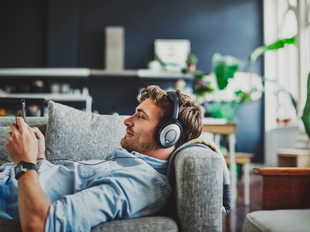 Мъж, отпускащ се на диван, слушащ музика със слушалки, с спокойно изражение на лицето